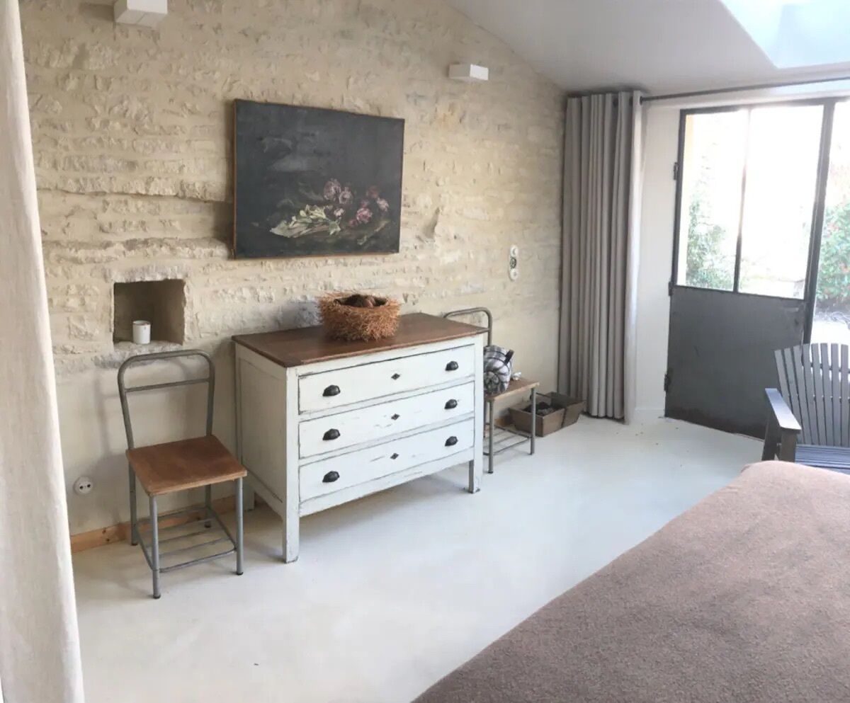 Een kamer met een rustieke bakstenen muur, een houten dressoir met lades, een metalen stoel en een groot raam met gordijnen.