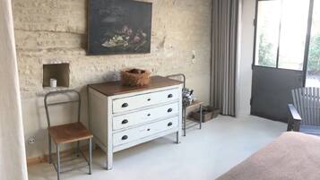 Een kamer met een rustieke bakstenen muur, een houten dressoir met lades, een metalen stoel en een groot raam met gordijnen.