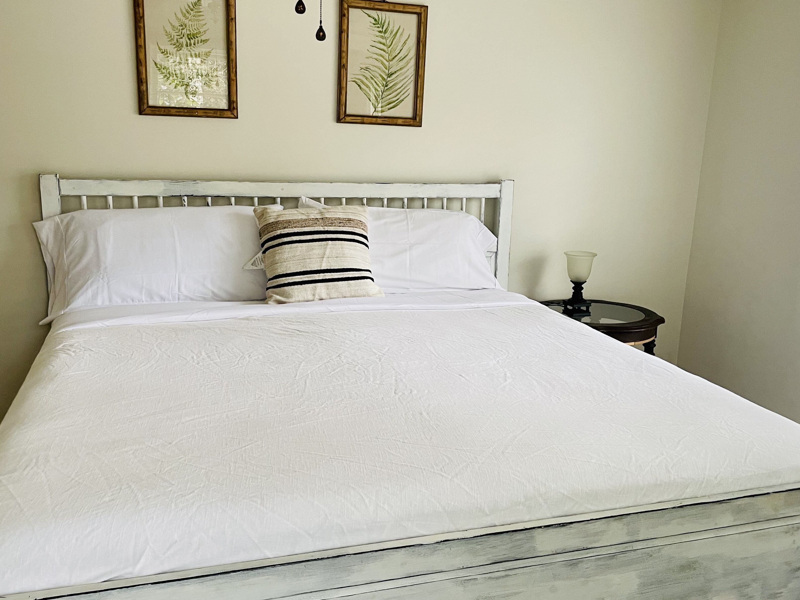 A neatly made bed with white linens, a striped pillow, and two framed botanical prints on the wall.
