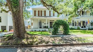 A white colonial-style building with a large tree in front, a well-maintained lawn, and a clear sky.