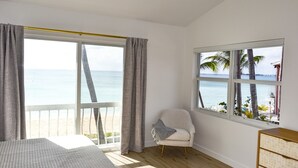 Quarto com uma grande janela com vista para uma praia, uma cama, uma cadeira e um penteoureiro.