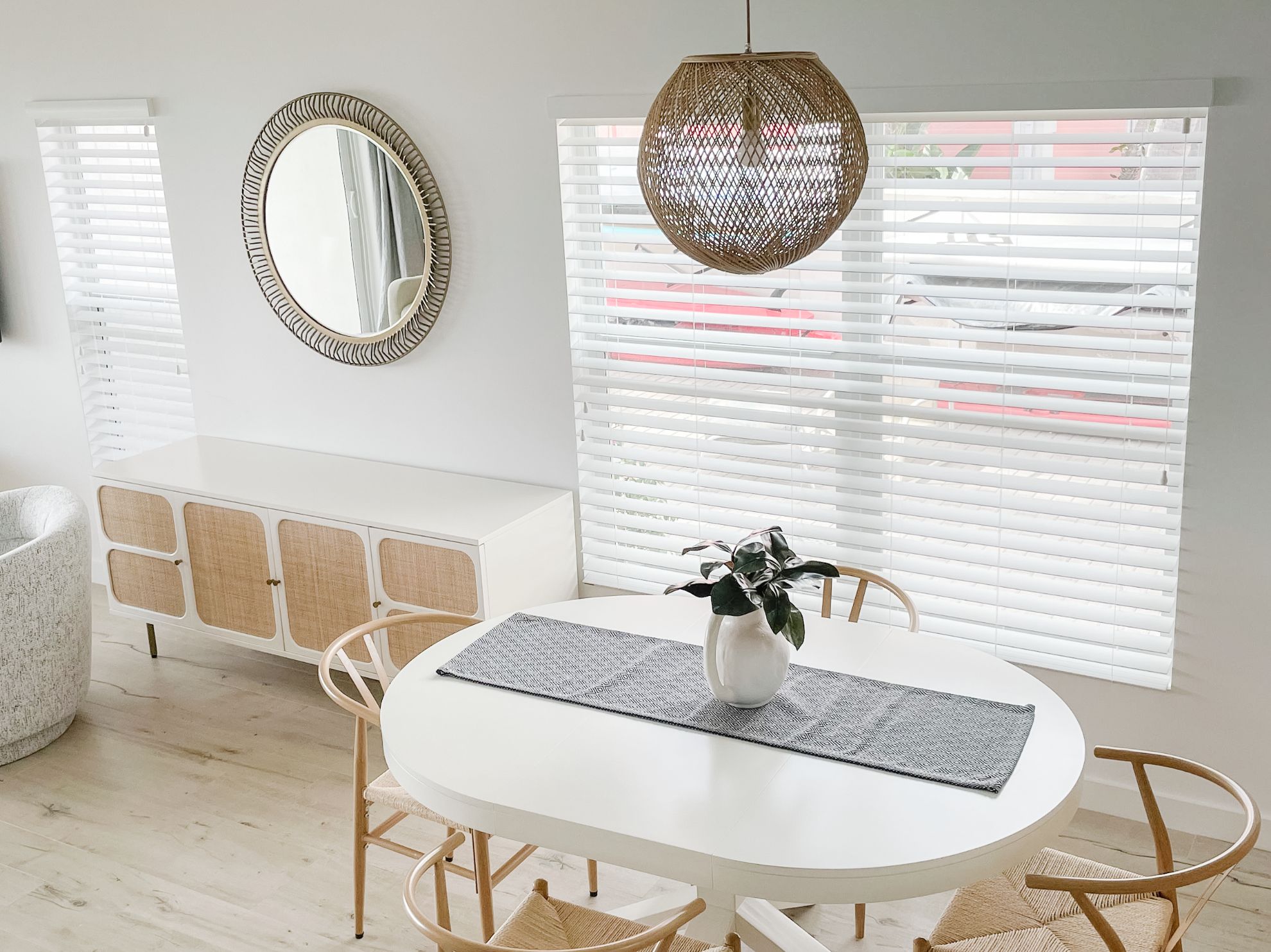 A dining area with a round white table, two chairs, a woven pendant light, a white vase with a plant, and a mirrored wall.