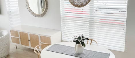 A dining area with a round white table, two chairs, a woven pendant light, a white vase with a plant, and a mirrored wall.