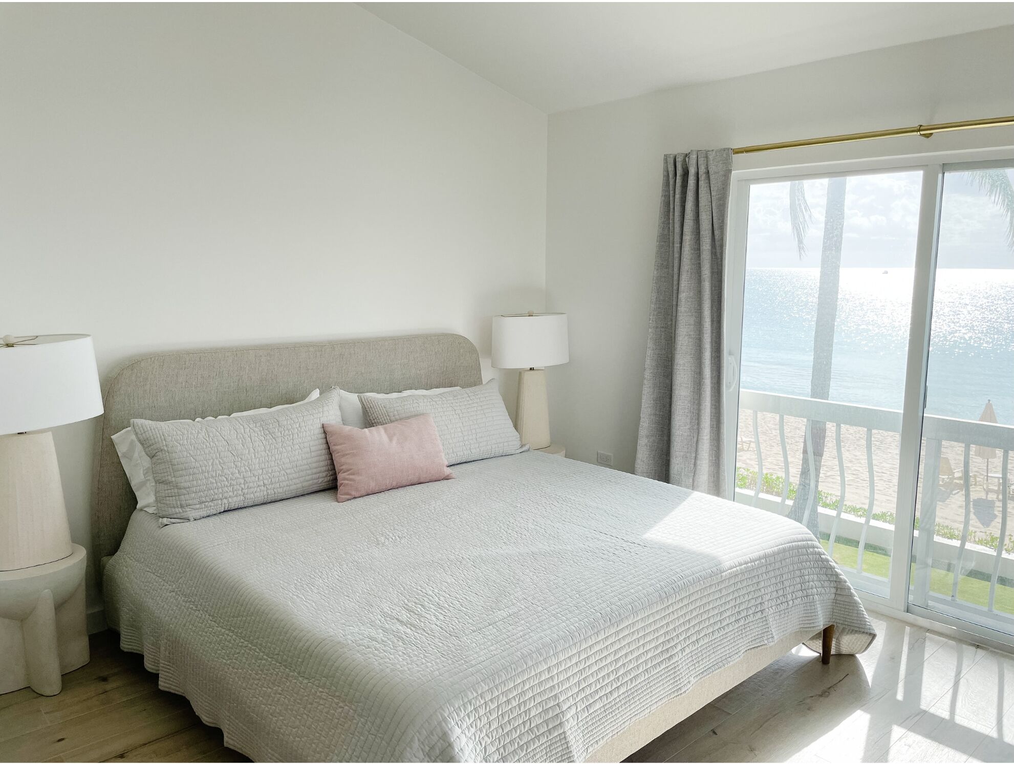 A bedroom with a bed, two bedside lamps, a view of the sea through a large window, and a balcony with a railing.