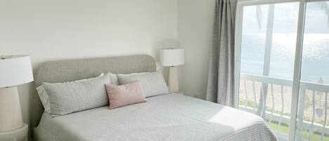 A bedroom with a bed, two bedside lamps, a view of the sea through a large window, and a balcony with a railing.