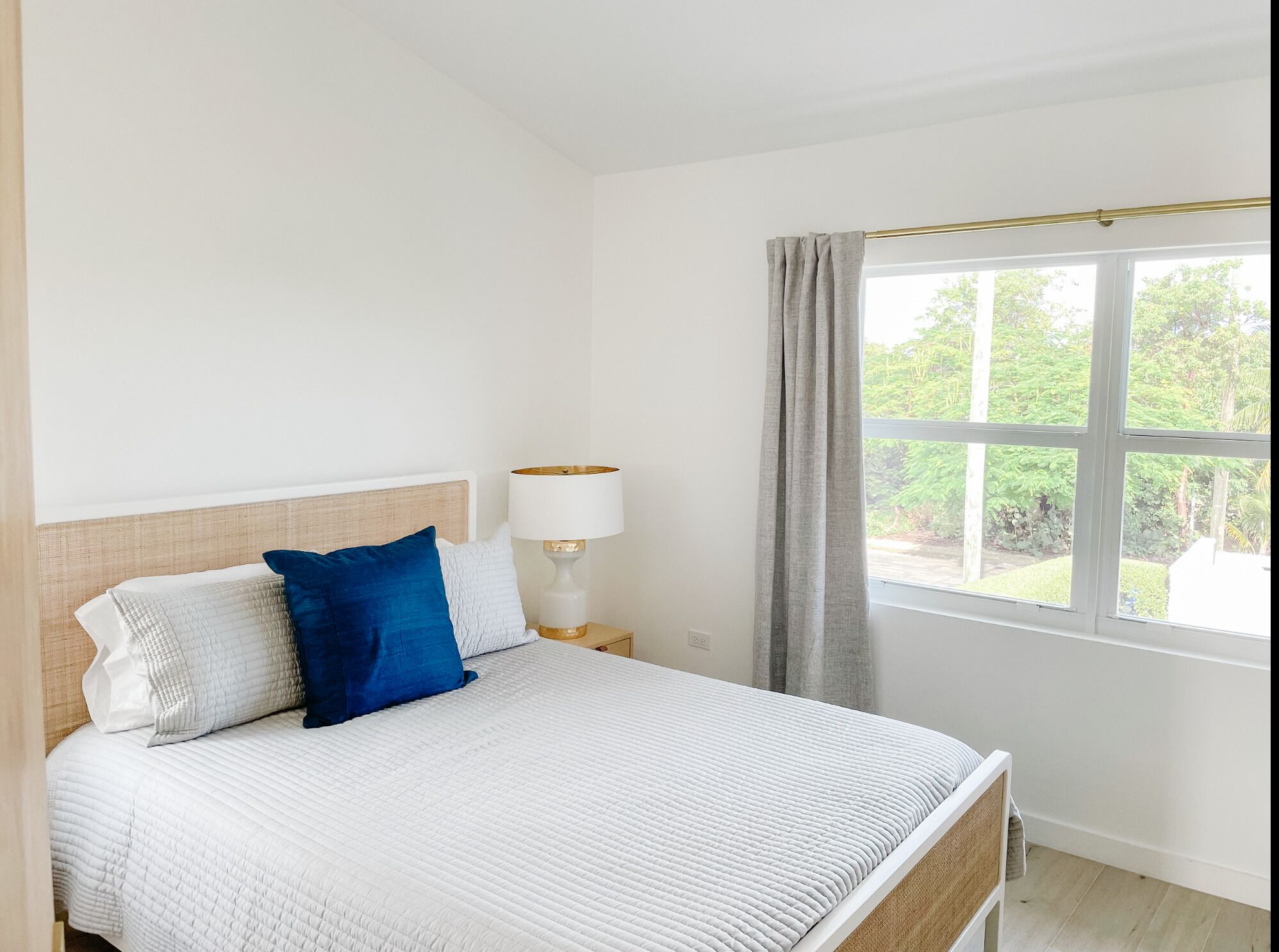 A bed with a white quilted bedspread, a wooden headboard, and two pillows (one blue, one white). A bedside lamp with a white shade and gold base is on the nightstand. A window with a view of greenery is visible.