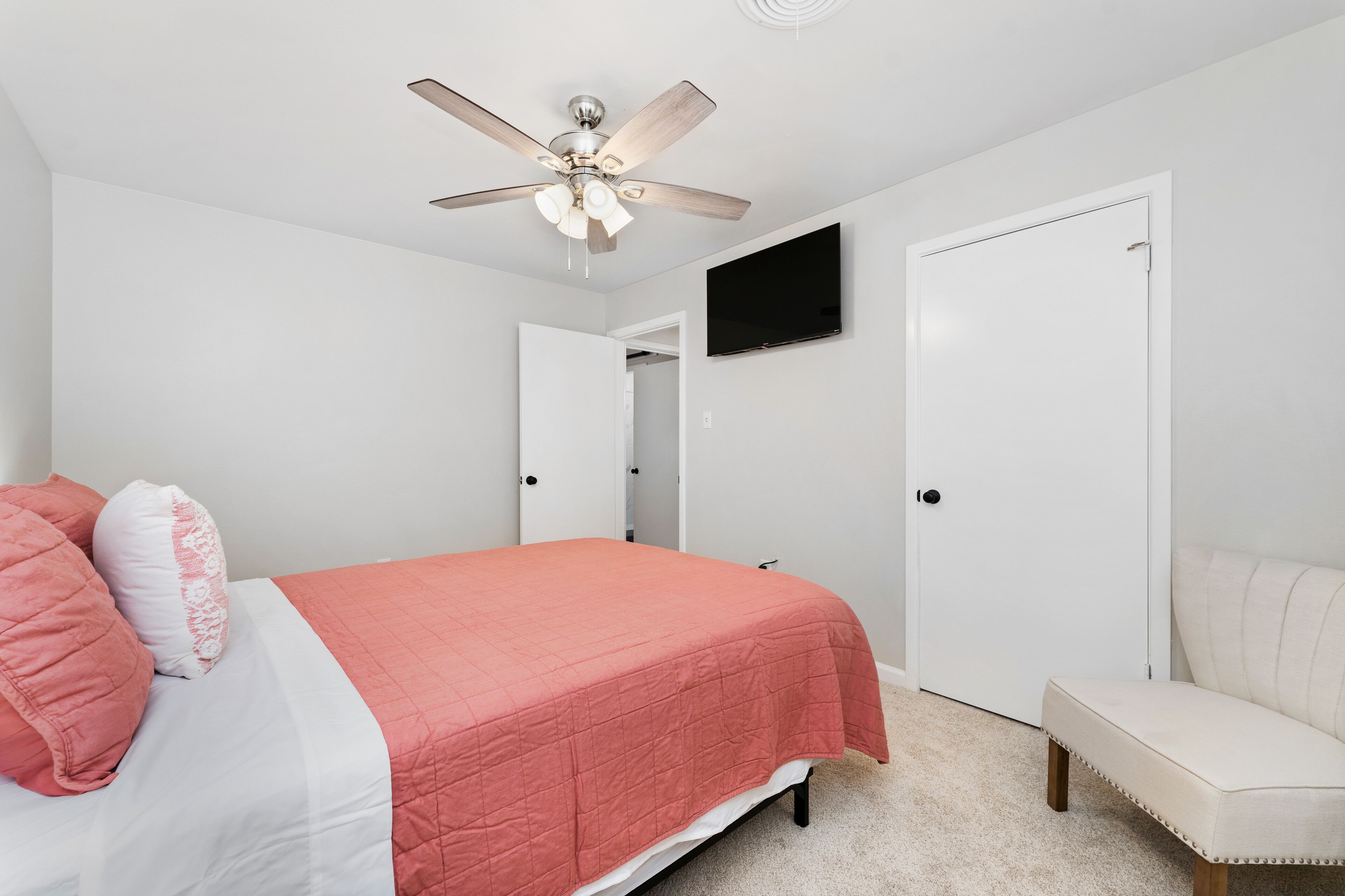 Un dormitorio con cama, ventilador de techo, televisor montado en la pared y una puerta.