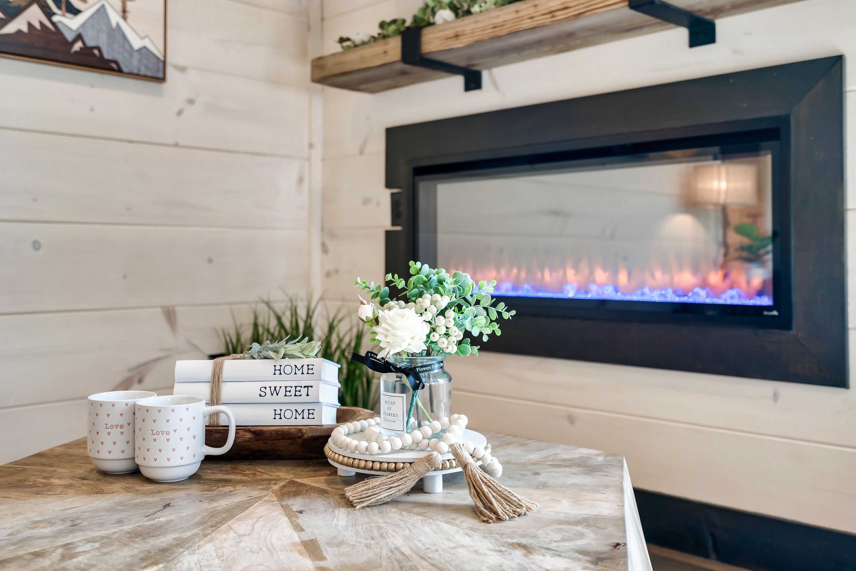 A cozy room with a fireplace, a table with decorative items, and a TV displaying a fire.