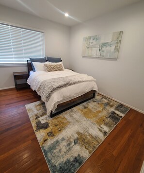A bedroom with a bed, a nightstand, a window with blinds, a wall art piece, and a patterned rug.