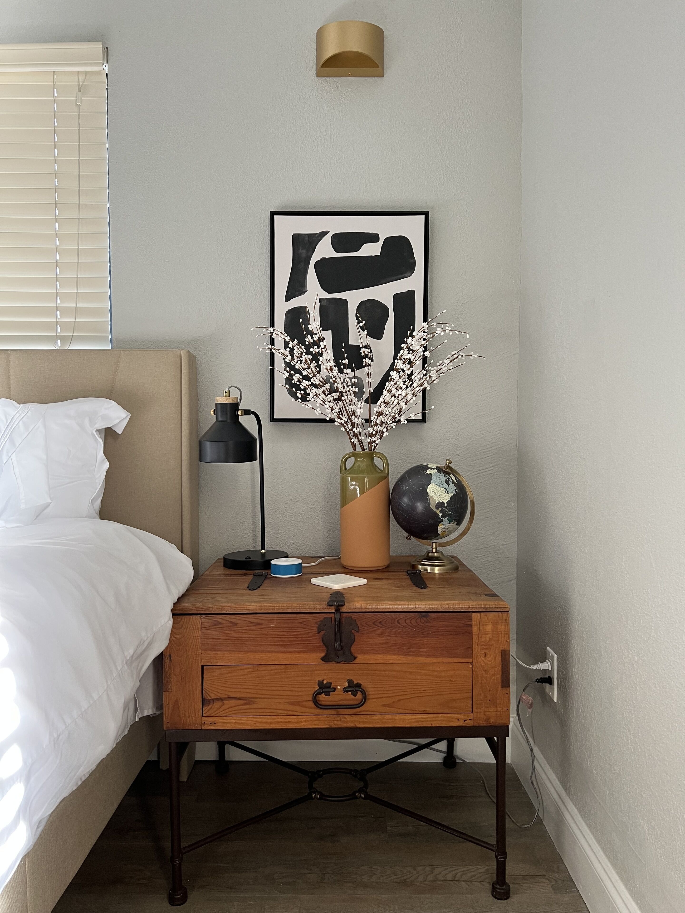 Un dormitorio ordenado con cama, mesita de noche, lámpara, un globo terráqueo y obras de arte.