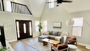 A spacious loft with a high ceiling, a black ceiling fan, a gray sectional sofa, a wooden coffee table, and a large window with blinds.