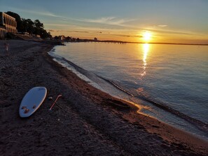 A beach with a surfboard and paddle on the sand, a pier in the background, and a sunset over the water.