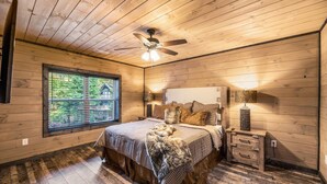 A cozy bedroom with a wooden ceiling, a large window, a bed with a checkered bedspread, and a nightstand with a lamp. - Great Location, 3 min To Parkway! Private Hot Tub,Arcade, Fireplace,shuffleboard (Pigeon Forge)
