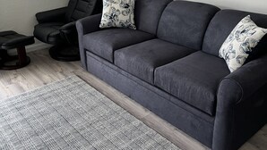 Ein Wohnzimmer mit einem dunkelgrauen Sofa, zwei gemusterten Zierkissen, einem gemusterten Teppich, einem schwarzen Sessel und einem Holztisch.