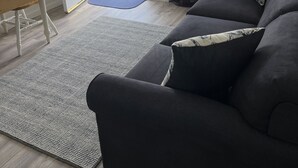 Ein Wohnzimmer mit einem dunkelgrauen Sofa, einem Holzstuhl, einem kleinen Schreibtisch, einem gemusterten Teppich und einer Tür mit einem Willkommensschild.