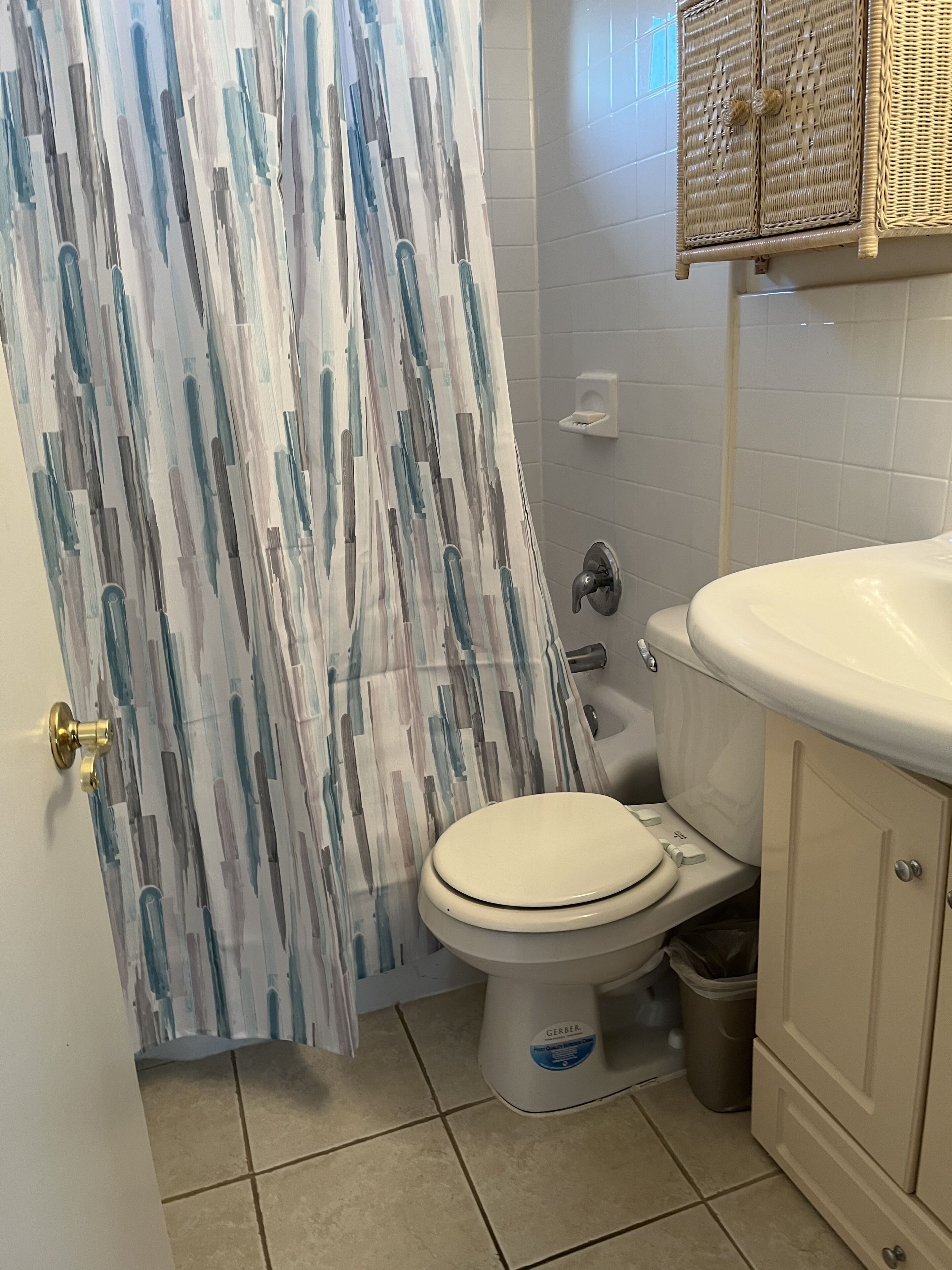 Un baño con inodoro, lavamanos, un cubo de basura y cortina de ducha con un diseño abstracto azul y blanco.