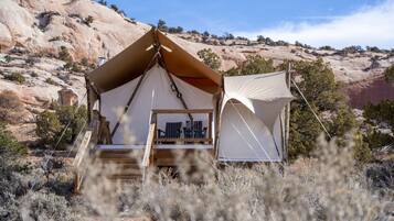 A tent with a wooden deck and a small table, surrounded by dry vegetation and a rocky landscape.