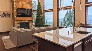 Une cuisine moderne avec une cheminée en pierre, de grandes fenêtres et une vue sur des arbres couverts de neige.