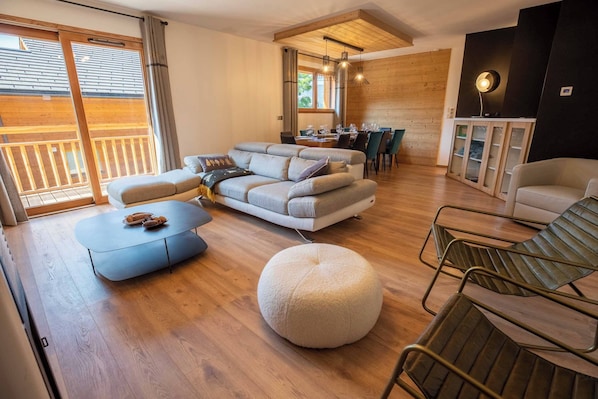A modern living room with a sofa set, ottoman, and metal chairs. A dining area is visible in the background with a table and chairs.