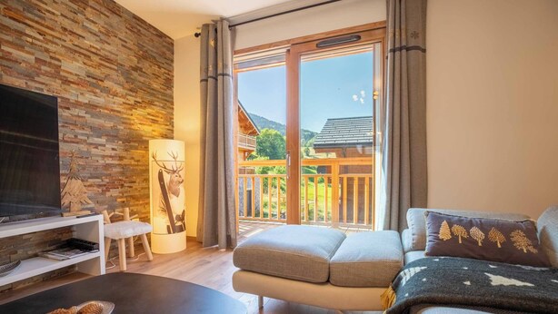 A modern living room with a flat-screen TV, a wooden wall, a sofa with cushions, and a balcony with a view of a forested area. - Chalet Gaïla spacious chalet in Les Allues next to the gondola (Meribel Les Allues)