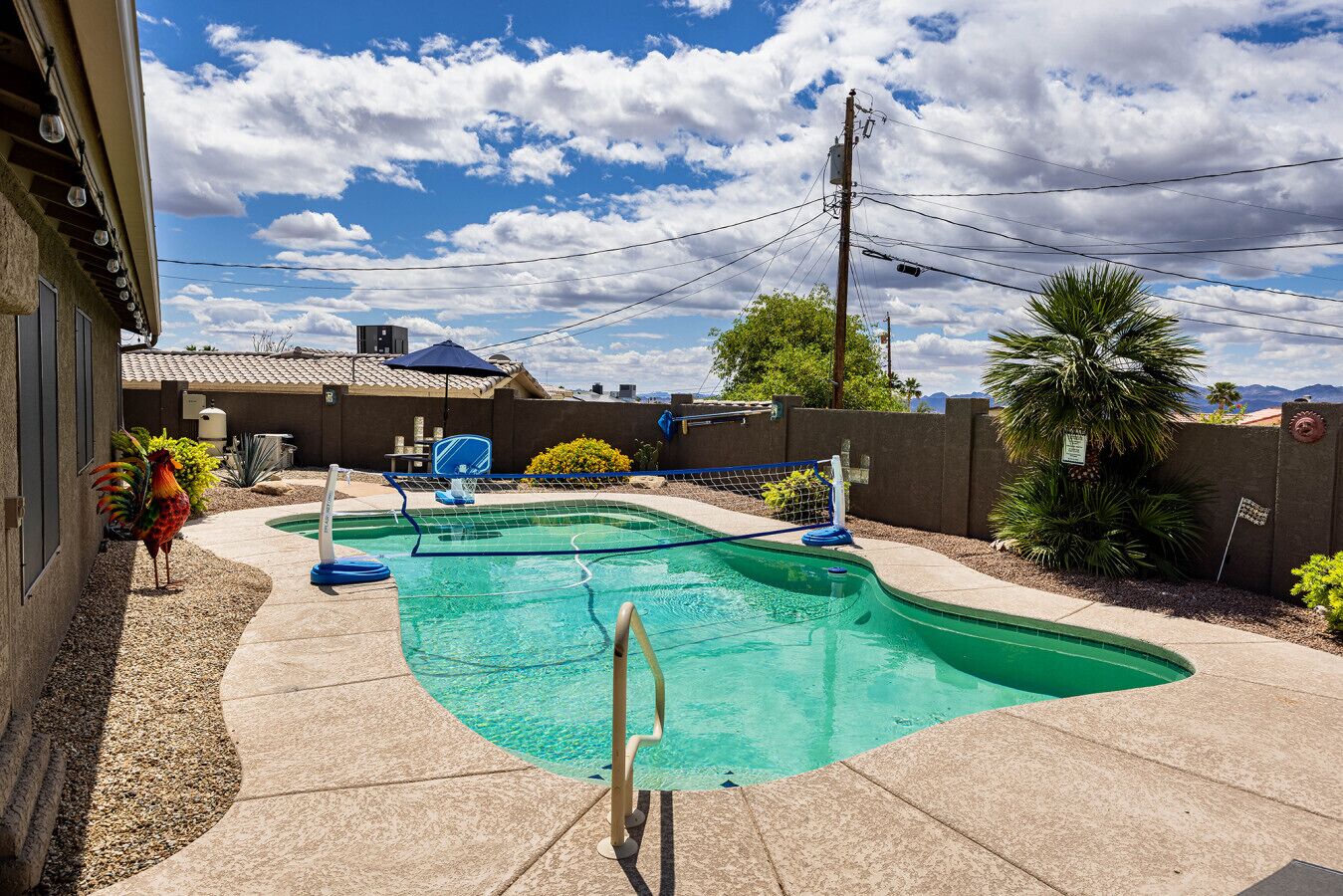 A swimming pool with a diving board, surrounded by a patio area with a fence, a palm tree, and a view of mountains.