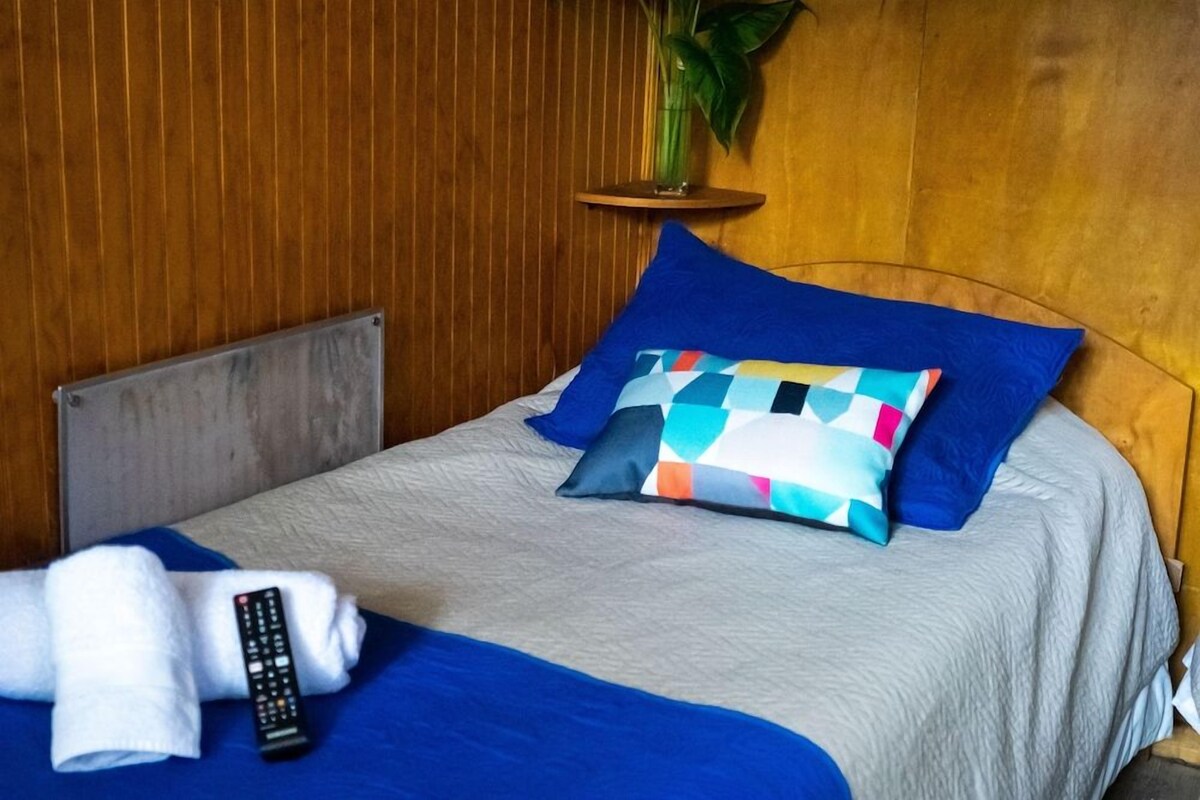 A neatly made bed with a blue and multicolored pillow, a remote control, and a white towel on the bed.