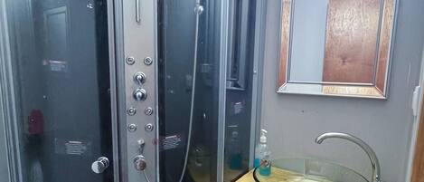 Un vestuario con ducha envuelta en vidrio, cabezal de ducha manual, tocador con lavabo de vidrio y espejo sobre el lavabo.