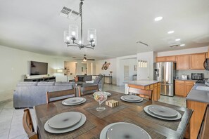 Una cocina moderna con electrodomésticos de acero inoxidable, un comedor con mesa de madera y manteles individuales grises, y una sala de estar con sofá gris y televisor de pantalla plana.