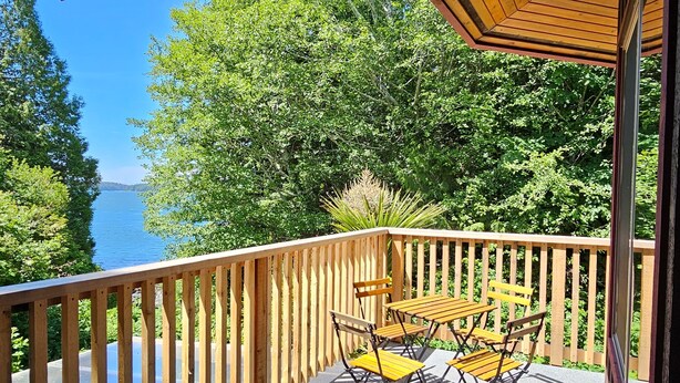 Terraza de madera con mesa y sillas, con vista a una masa de agua rodeada de árboles.
