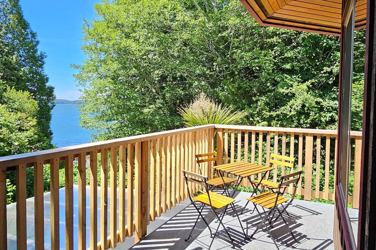 Een houten terras met een tafel en stoelen, met uitzicht op een waterpartij, omringd door bomen.