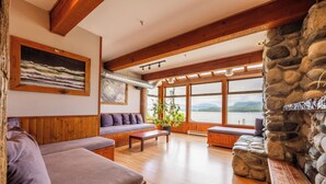 Una habitación amplia con suelos de madera, chimenea de piedra, ventanas grandes con vistas a un lago y techo de madera.