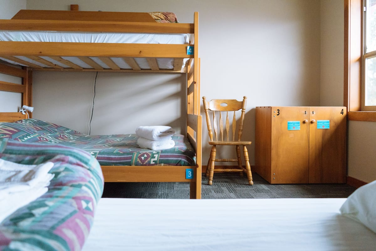Een stapelbed met een houten hoofdbord, een enkel bed met een geborduurd dekbed, een houten stoel en een kledingkast met genummerde deuren.