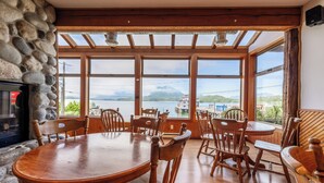 Un comedor con pared de piedra, sillas de madera, una mesa de madera y ventanas amplias con vistas a un lago y montañas.