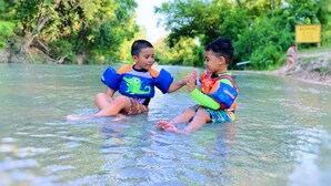 Zwei Kinder mit Schwimmwesten in einem flachen Fluss, umgeben von viel Grün.