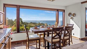 Un comedor con una mesa dispuesta para cuatro, ventanas amplias que ofrecen vistas al océano y al follaje, y una lámpara colgante moderna.