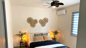 A bedroom with a bed, two bedside tables, a ceiling fan, wall-mounted air conditioning, and a window with blinds.