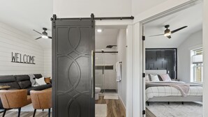 A modern bedroom with a bed, a sliding door, a bathroom, and a living area with a sofa and chairs. - NEW!!! Modern cottage and perfect location! (Bluffton)