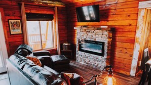 Interior acogedor de una cabaña con un sofá de cuero, una chimenea de piedra y un televisor montado en la pared.
