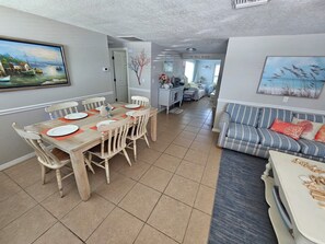 A dining area with a wooden table and chairs, a kitchen in the background, and a living area with a sofa.