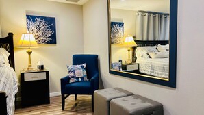 Quarto de hotel com cama, uma cadeira, mesa de cabeceira, abajur, espelho e um banco.