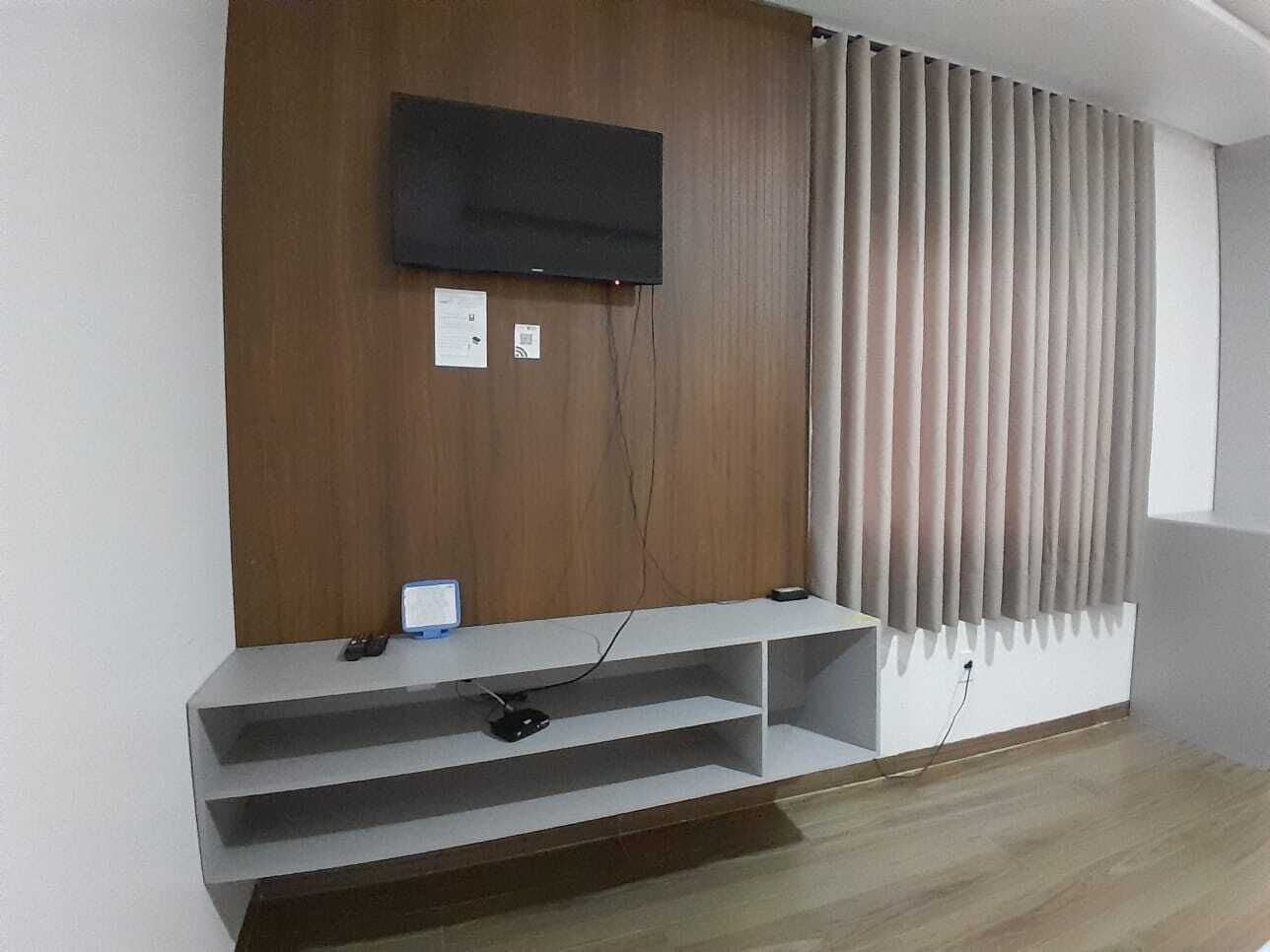 A room with a mounted TV, a wooden panel wall, a window with blinds, and a built-in shelf with a remote control.