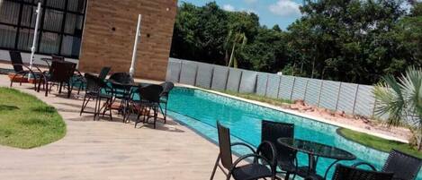 Un área de piscina moderna al aire libre con un edificio de madera, sillas de descanso y un cielo azul despejado.
