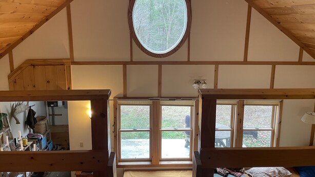 Una habitación con techo de madera inclinado, un tragaluz circular grande y vista de una persona al otro lado de la ventana.
