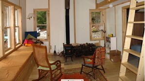 Interior acogedor de una cabaña con una estufa de leña en el centro, ventanas amplias y una zona en el entrepiso.