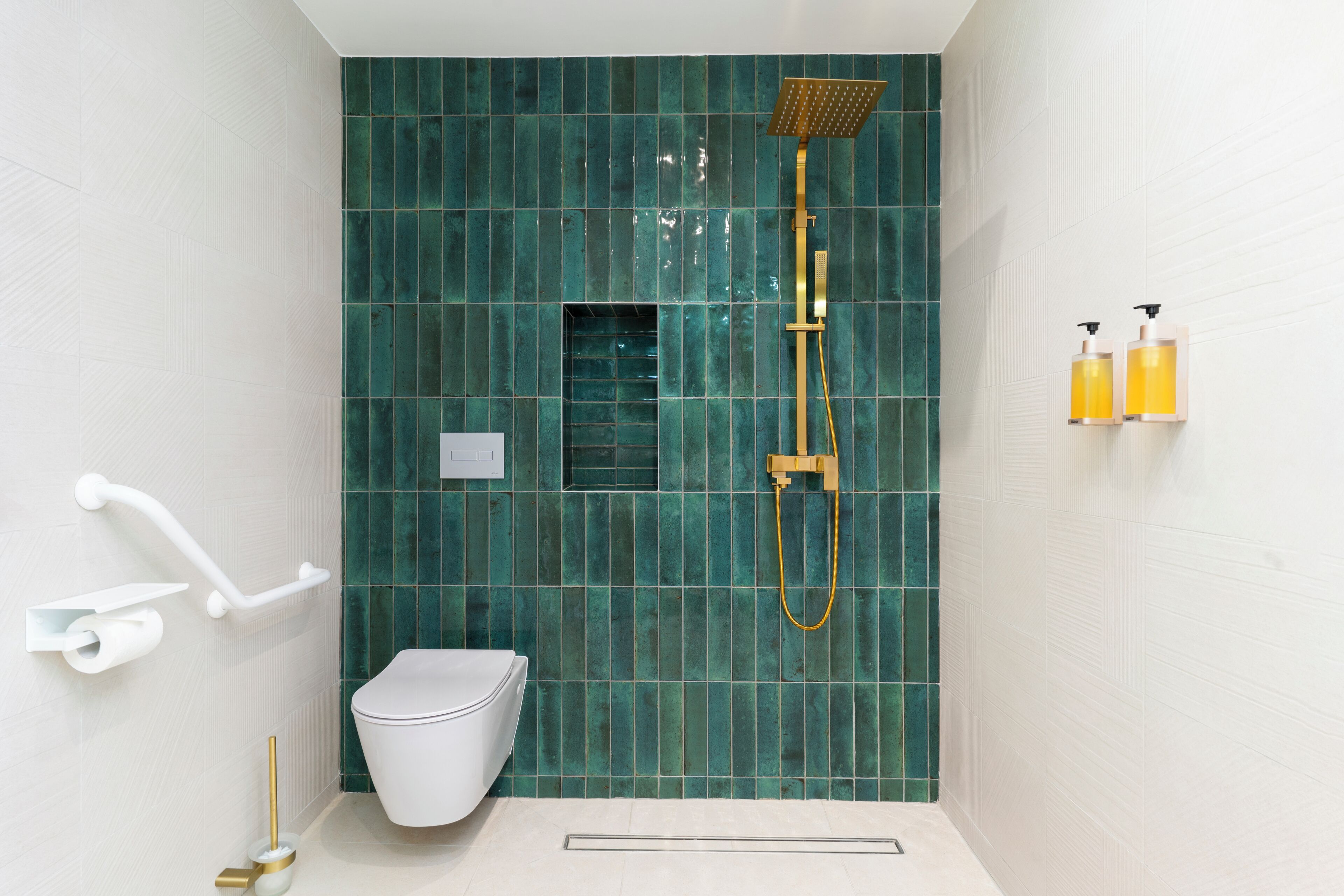 Une salle de bain avec des toilettes blanches, une douche dotée d’un pommeau de douche doré et deux bouteilles jaunes accrochées au mur.