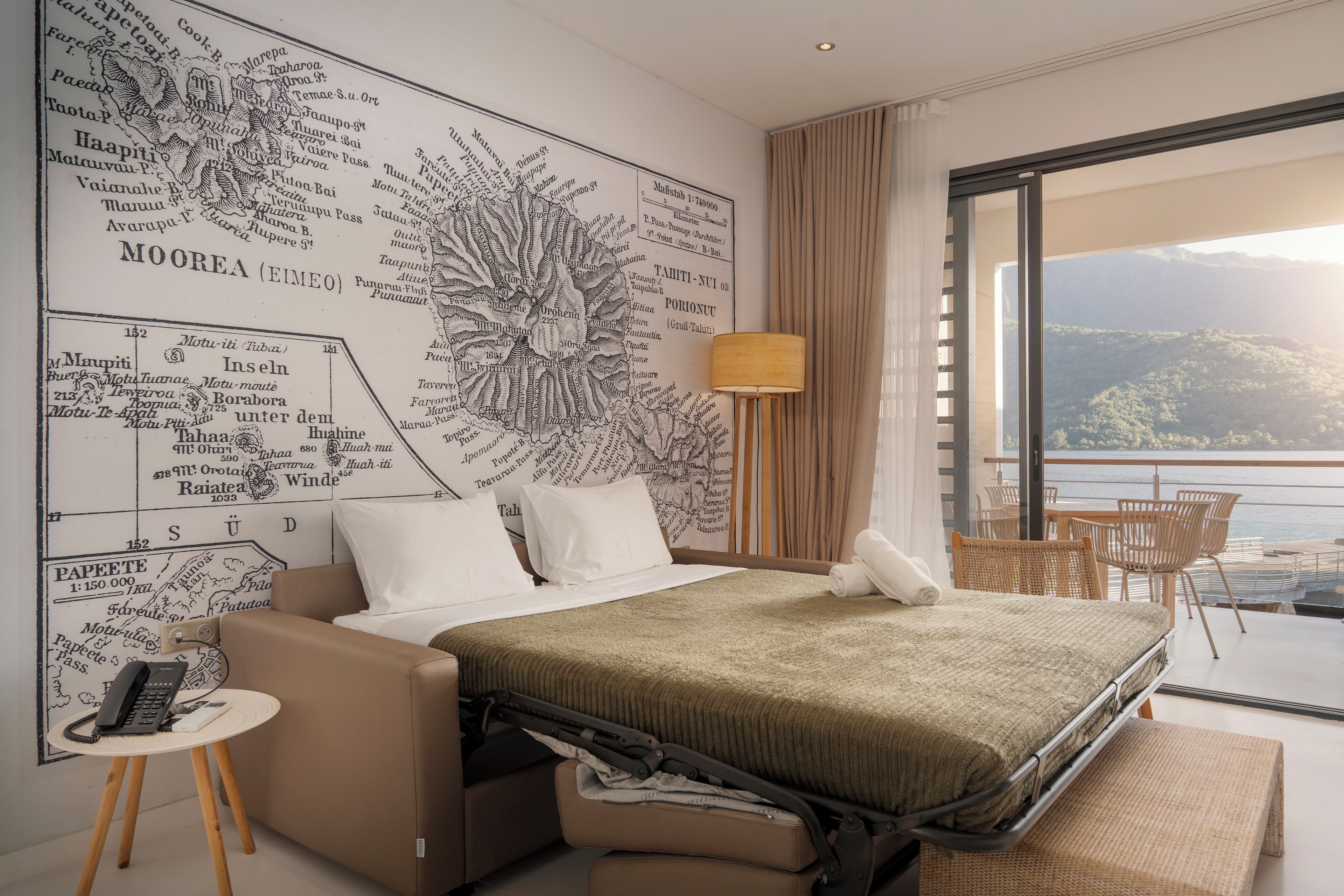 A hotel room with a large map on the wall, a bed, a small table with a phone, and a balcony with a view of the mountains.