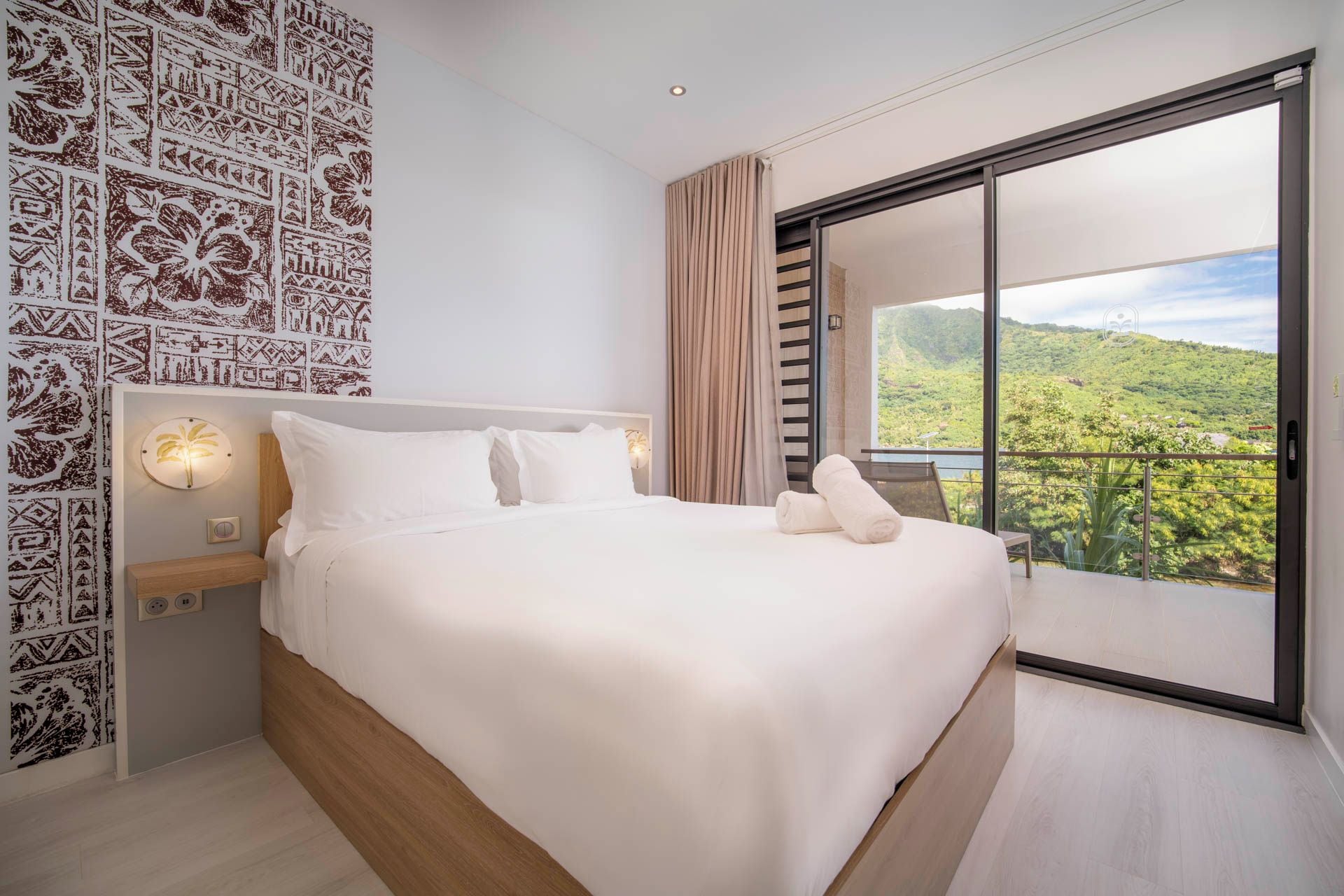 A modern bedroom with a large bed, a nightstand, a wall-mounted light, and a view of a lush landscape through a sliding glass door.
