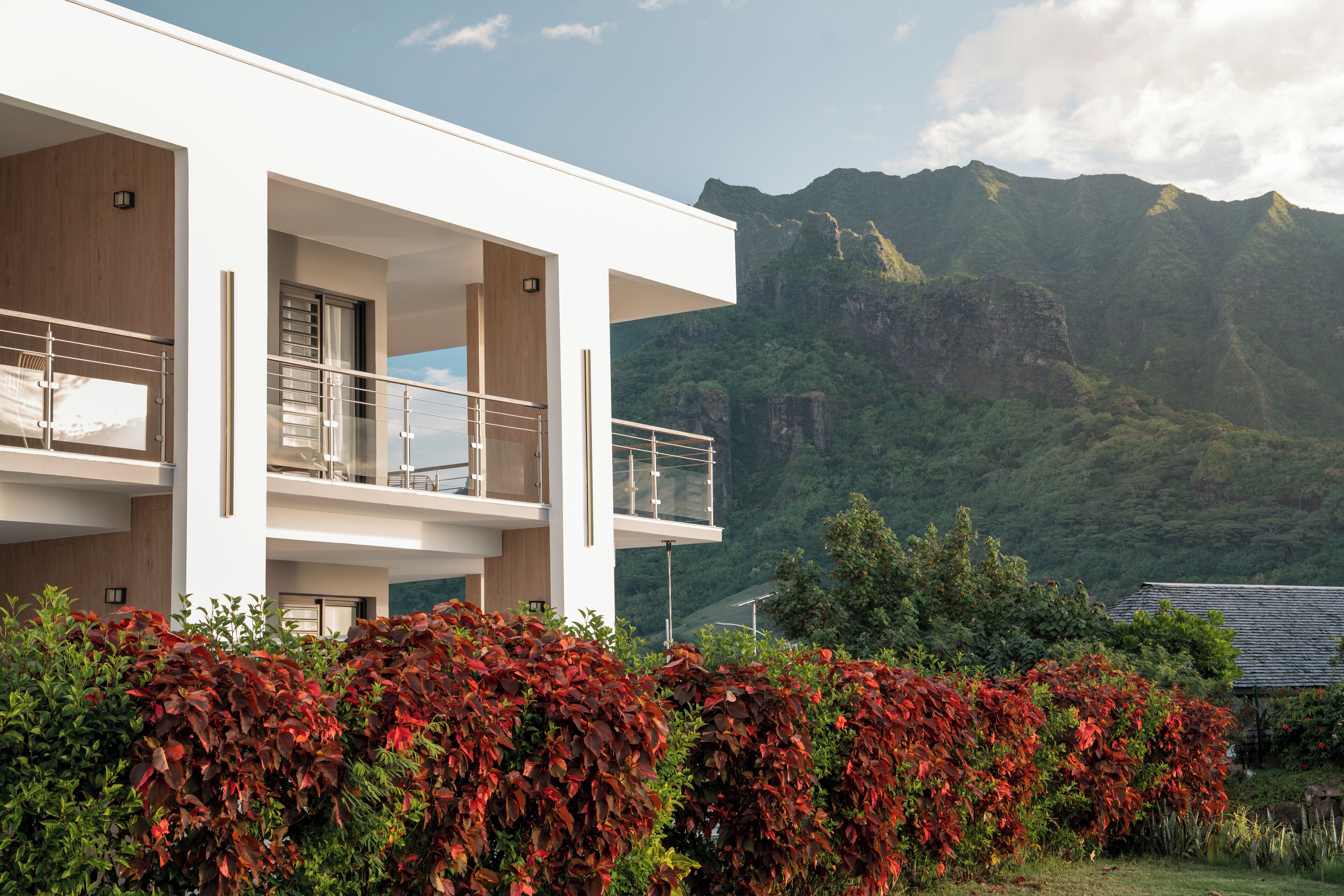A modern two-story building with a balcony, surrounded by lush greenery and mountains in the background.