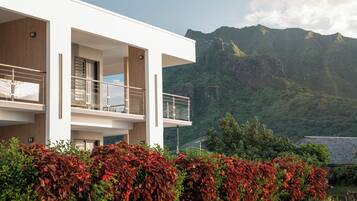 A modern two-story building with a balcony, surrounded by lush greenery and mountains in the background.