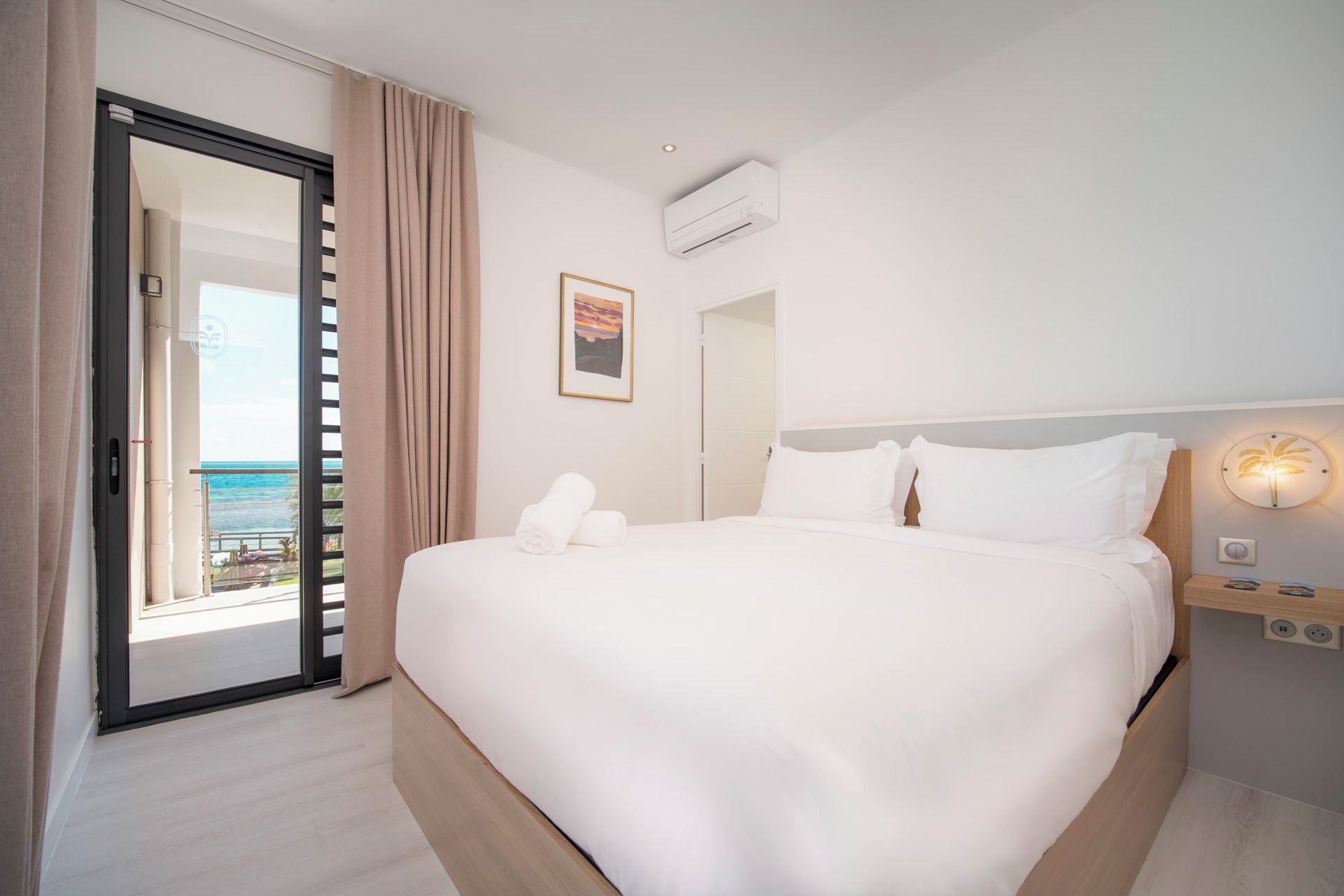 Habitación de hotel con cama, mesita de noche, equipo de aire acondicionado, luz empotrada en la pared y vista al mar a través de una puerta corrediza.